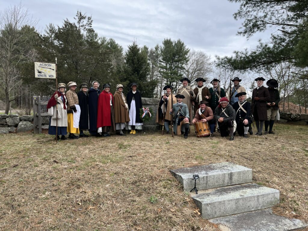 A group of men and women in revolutionary era garb. Many of the men are carrying muskets. The women have an Acton Minute Men flag.