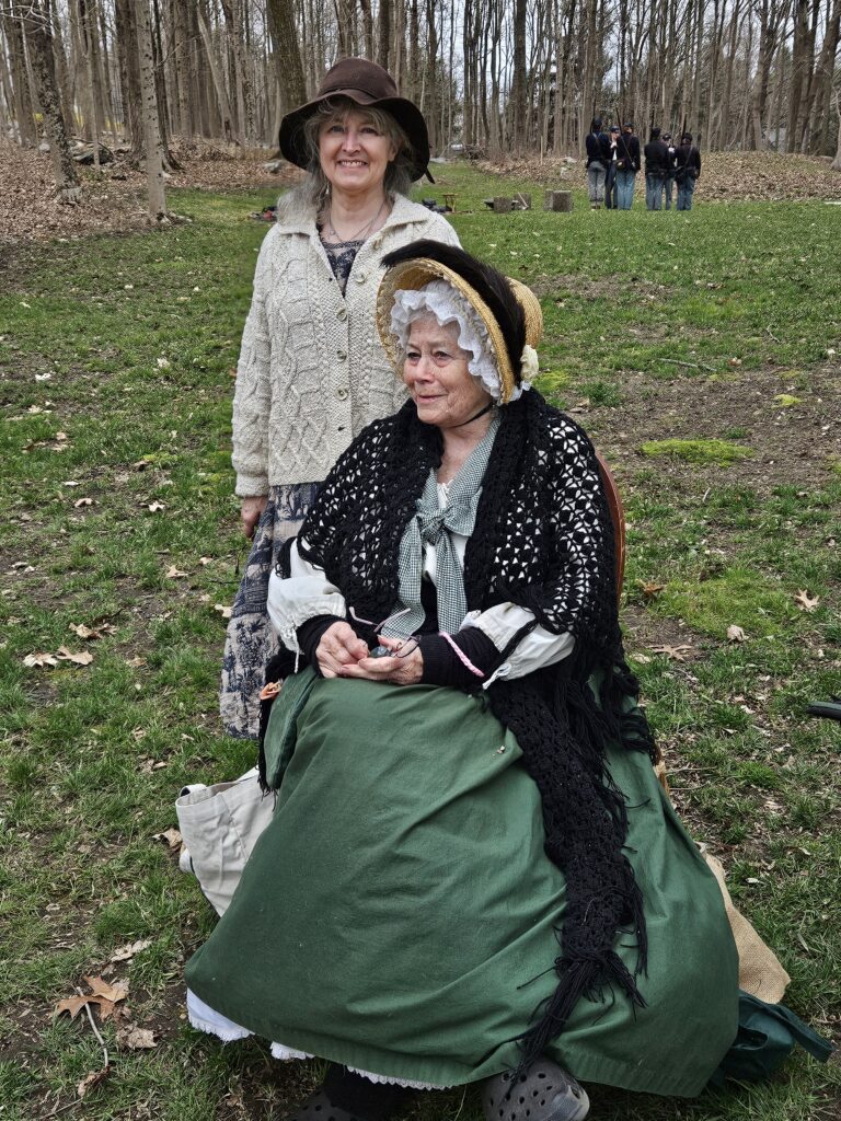 Two women , one wearing 18th century garb, complete with a mob cap and straw hat.