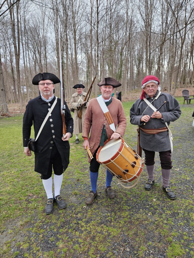 Four men in revolutionary era garb. Two men are carrying muskets, one has a drum and one has a flute.