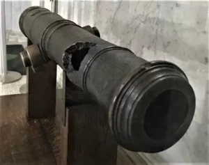 A black cannon on a stand. The cannon has a large, ragged hole.