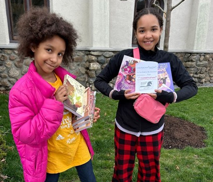 Two girls wearing bright clothing with armfuls of books and big smiles.The girl on the right is showing her award for her cover illustration.