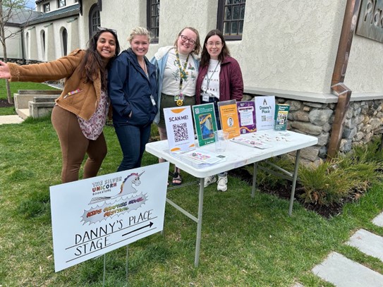 Four women stand behind a table with information about the festival and Danny’s Place. There’s a sign pointing to the Danny’s Place stage.