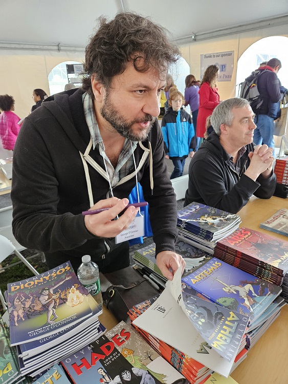 A bearded man with curly hair holds a pen in one hand and book in the other. He is talking intently with whoever is waiting for his autograph.