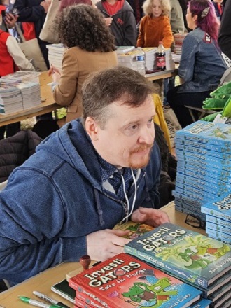 Man with a goatee and blue sweatshirt sits with a stack of his Investigators graphic novels.