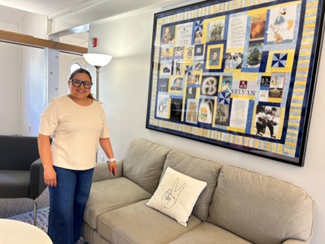 A woman with blonde hair and big glasses stands next to a framed quilt hanging on a wall. The quilt includes pictures of Danny McCarthy and mementos of his life.