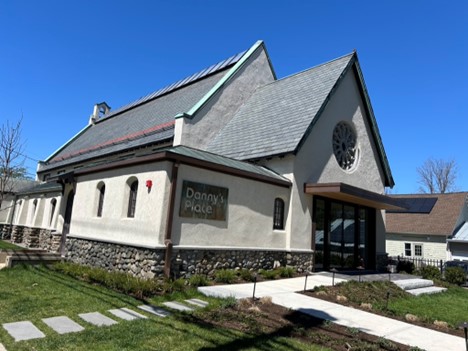 A stucco and stone building with a steep roof, a rose window and the Danny's Place sign on the front.