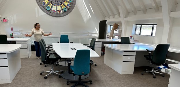 A woman shows off a well-lit room with lots of desks that are large enough for art projects or for people to work together. There's a beautiful stained glass rose window on the wall.