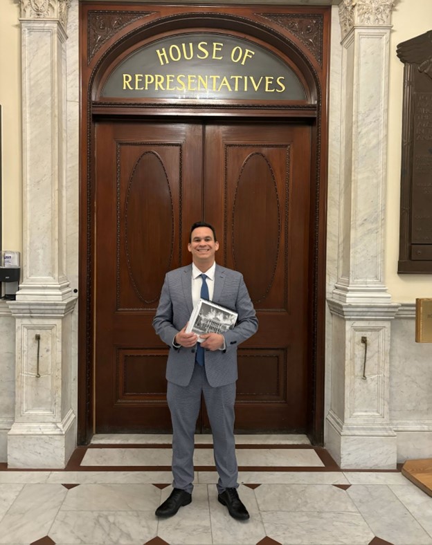 Man wearing gray suit stands in front of a door labeled House of Representatives. He is smiling and holding a copy of the MA 2025 budget in his hands.