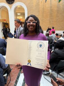A woman with black hair, big pink glasses, and wearing a purple dress holds an award (the Black Excellence on the Hill Award).