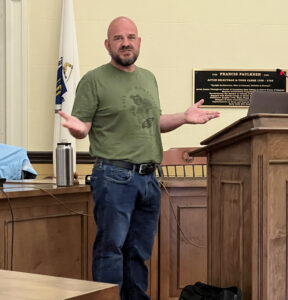 A bald, bearded, man stands in front of a podium. He is wearing jeans and a t-shirt, as befits a gardening expert.