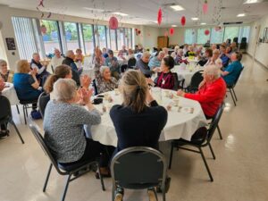 A decorated room with several tables full of people. At the front table, a woman raises her hand.