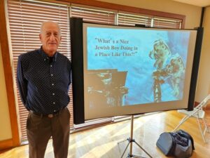 A tall man stands next to a screen. The screen shows a picture of two astronauts on a space walk. The image is labeled "What's a Nice Jewish Boy Doing in a Place Like This?"