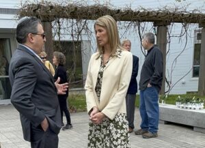 A man wearing a suit and a yarmulke talks with a very tall woman wearing a jacket and flowered dress. They are on the patio of a building (Congregation Beth Elohim) and there is a trellis in the background.
