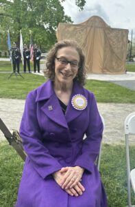 A smiling woman in a purple coat sits in a chair outdoors. She is wearing a large purple and gold ribbon rosette that says "Meredith Bergmann, Sculptor."