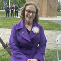 A smiling woman in a purple coat sits in a chair outdoors. She is wearing a large purple and gold ribbon rosette that says "Meredith Bergmann, Sculptor."