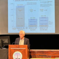 A grey-haired man in a suit jacket stands at a podium. A large screen is behind him with a page from his presentation on leaf blowers.