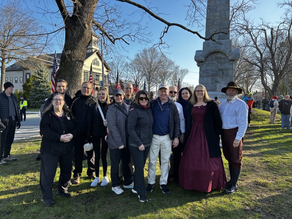 A group of people of various ages, some in street clothes and some in costume, stand in front of the monument in Acton.