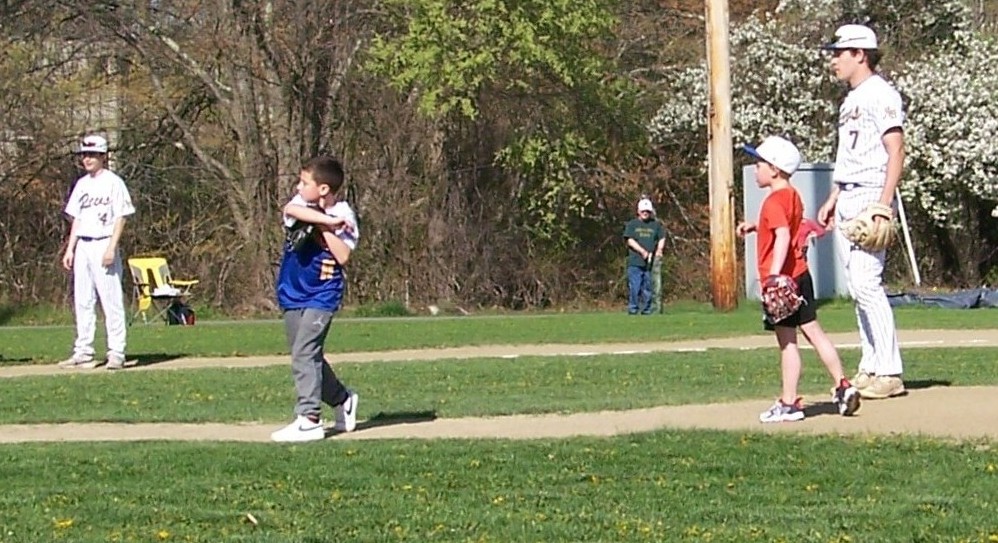 Two grade school boys stand with two High School players. One of the boys is throwing a ball (inelegantly).
