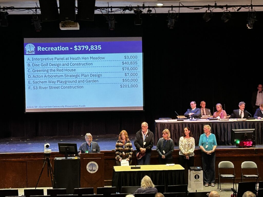 A long shot of a large screen (with CPC requests). In front of the stage, 6 people are lined up. One is speaking at a mic. Behind him is the screen, and to the right are a number of men and women seated at a table.