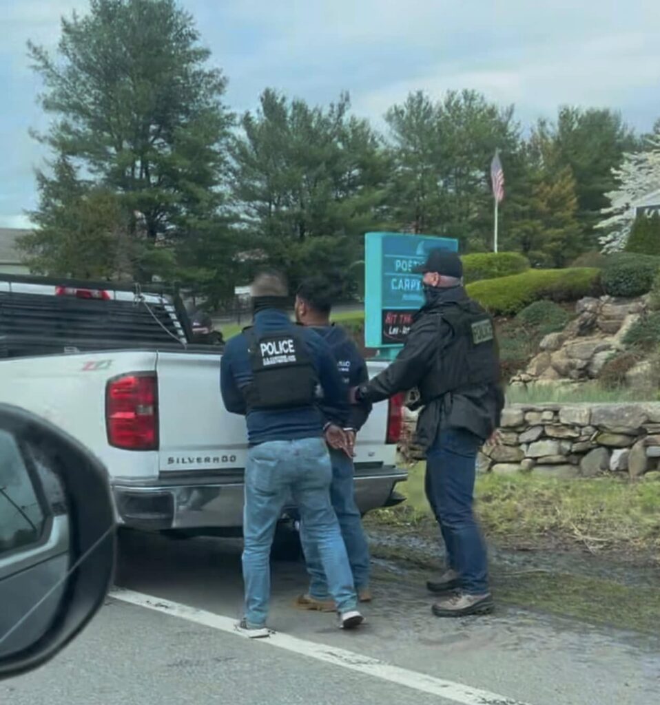 Three men standing behind a white pickup truck. Two men are wearing protective vests labeled Police and black neck gaiters pulled up over their faces, and are holding the arms of the third man. The third man has his wrists shackled behind him.