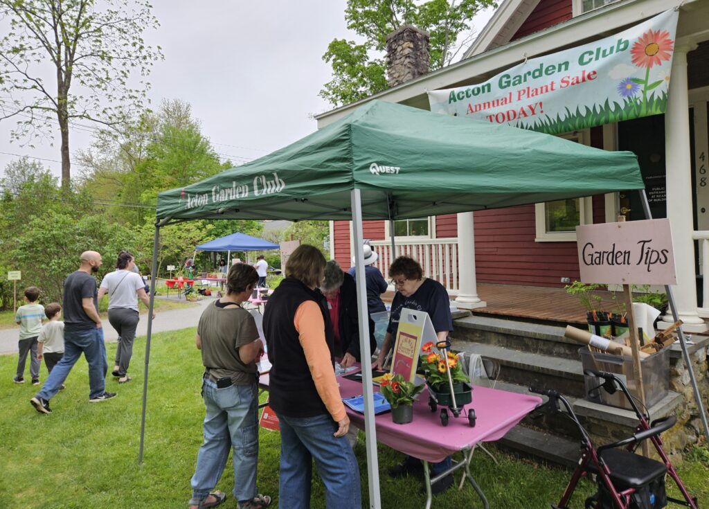 Inder a green tent with a Garden Tips sign, one woman stands behind the table and several peple are asking questions.