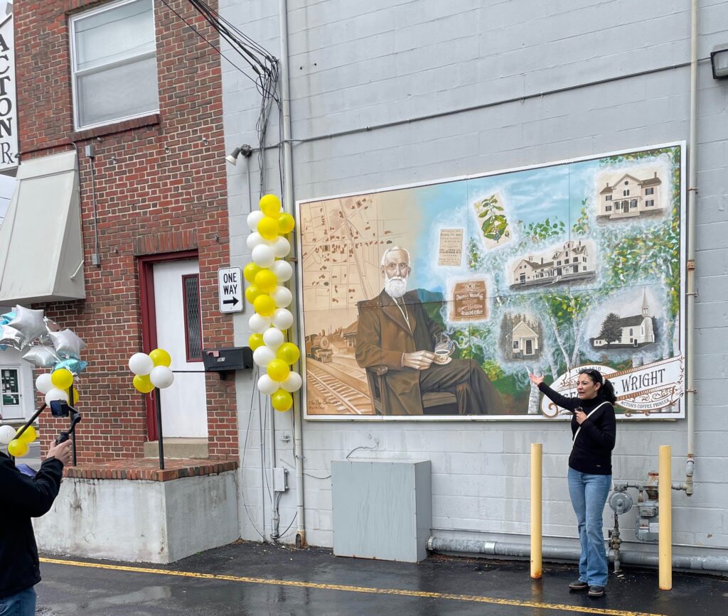 A womanholding a mic stands in front of the mural. The mural features an older man sitting with a cup of coffee on his knee, a map of early Acton and a picture of the railroad is on the left, and pictures of several buildings are on the right.