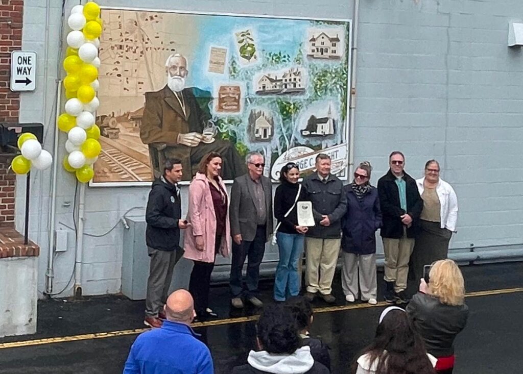 A group of people stand in front of the mural. A artist, wearing black, holds a citation.