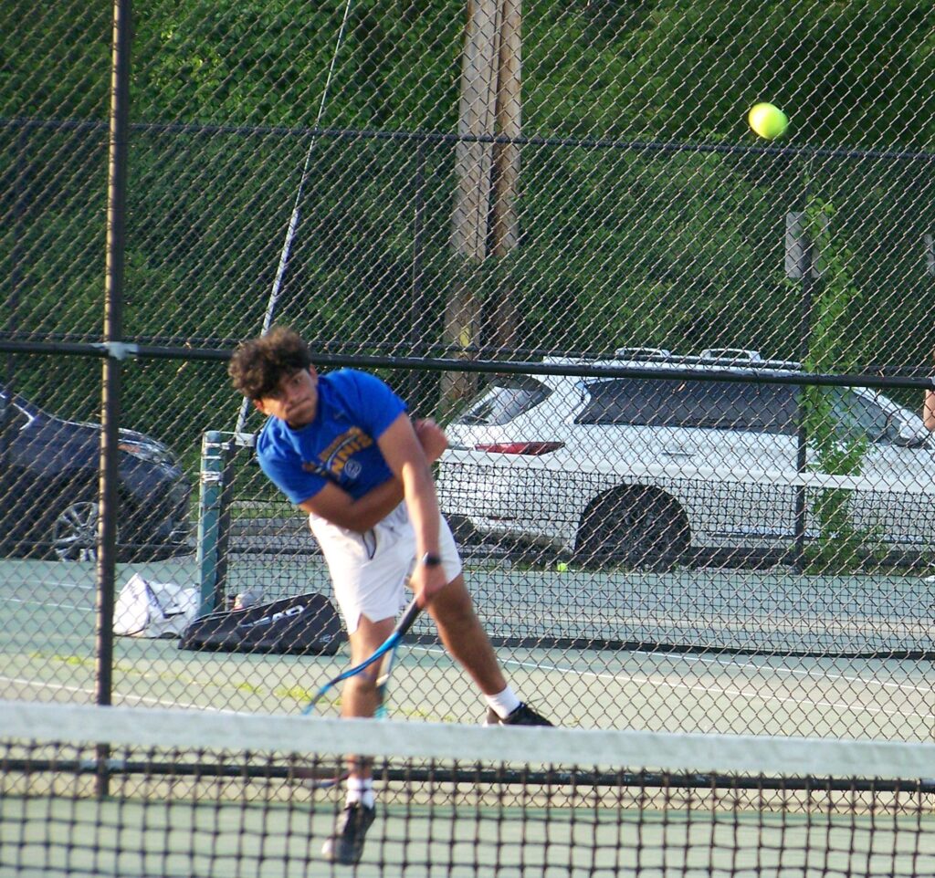 An AB tennis player has just hit finished and underhand stroke. The yellow ball is sailing towards the net.