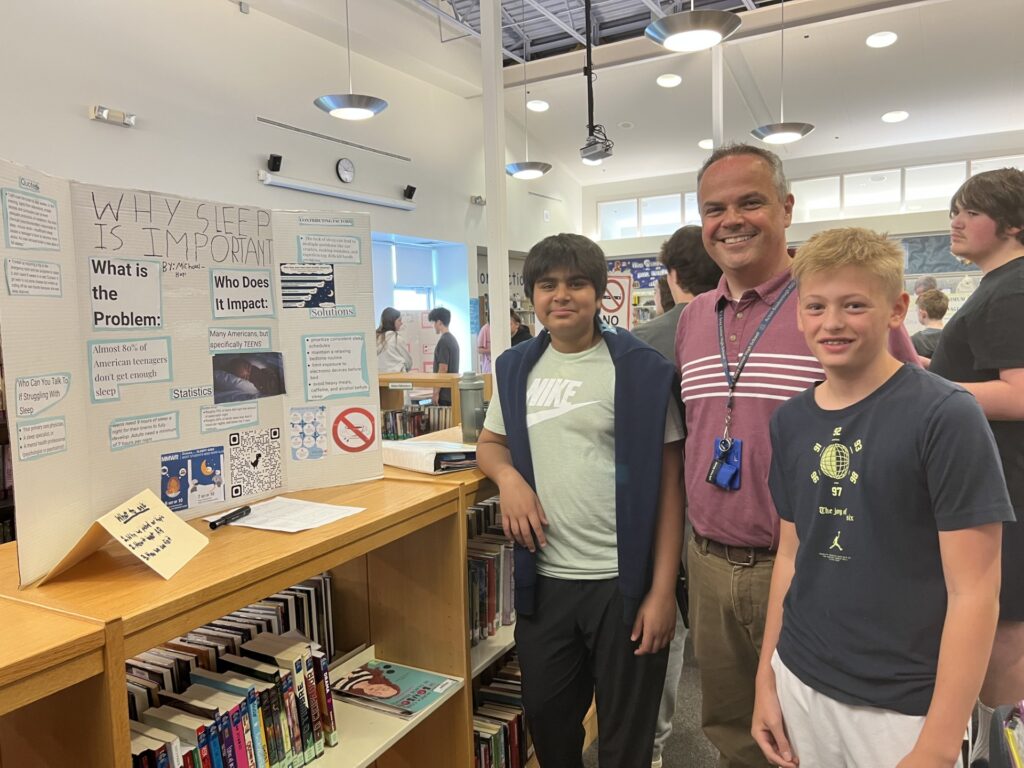 A man wearing a striped polo shirt stands with two boys next to a poster that is titled "Why Sleep is Important."