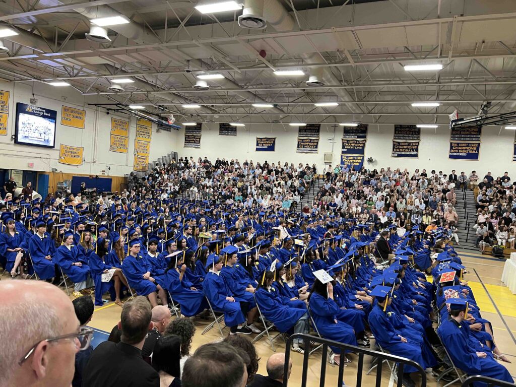 Students in blue gowns sit quietly. The bleachers in the background are full of spectators (mostly parents and guardians).