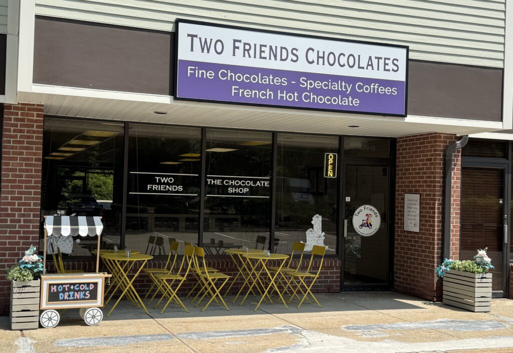 A store front with a large sign that says "Two Friends Chocolates: Fine Chocolates - Specialty Coffees - French Hot Chocolate. Underneath, a few yellow cafe tables and chairs await customers.