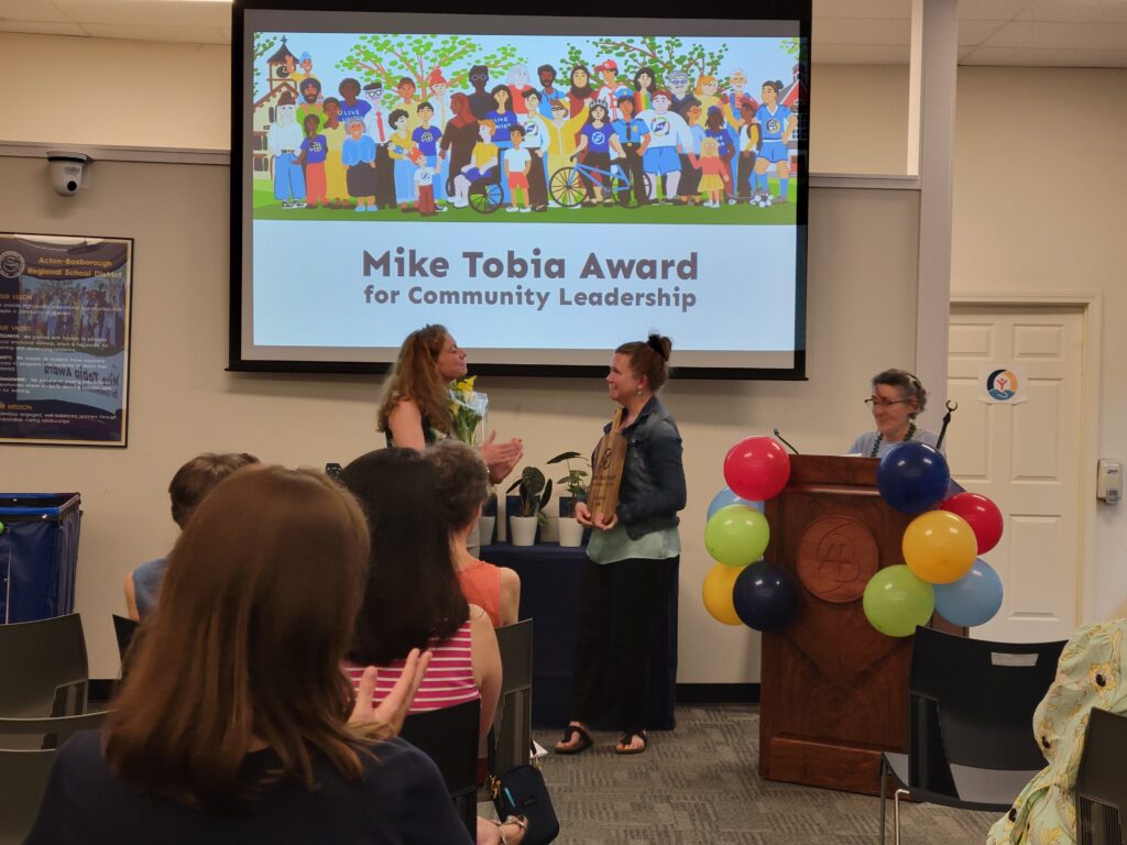 A woman in a long black skirt stands under a screen that proclaims "Mike Tobia Award for Community Leadership." On the left, a woman in a black dress holds flowers, and on the right, a woman speaks behind a balloon-covered podium.