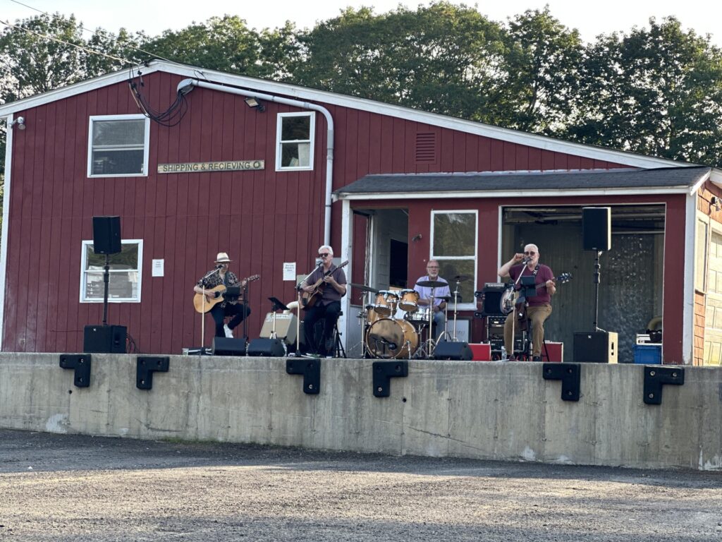 A band (two guitars, drums, and bass play on a concrete platform in front of a red wooden building.