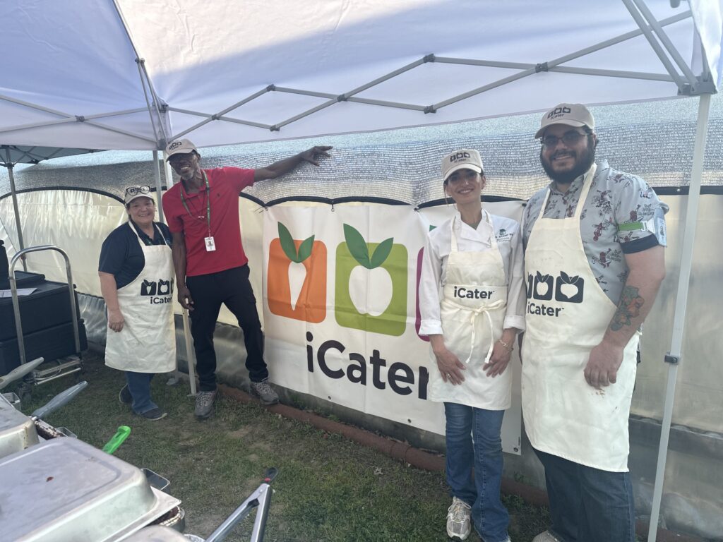 Three people wearing iCater aprons and a large man in a red polo shirt stand under a tent. The back wall of the tent also has the iCater logo.