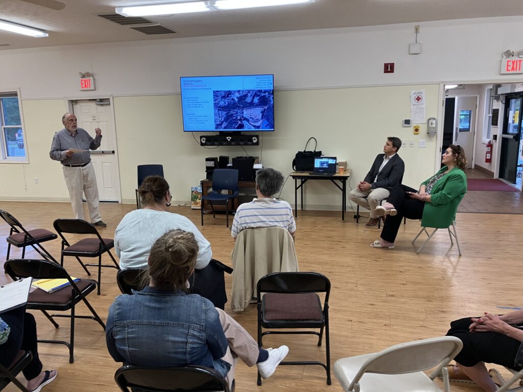 A man stands rear a a screen that contains a picture and text. The Town Manager and Economic Development Director sit to the right of the screen and people sit in the audience in the foreground.