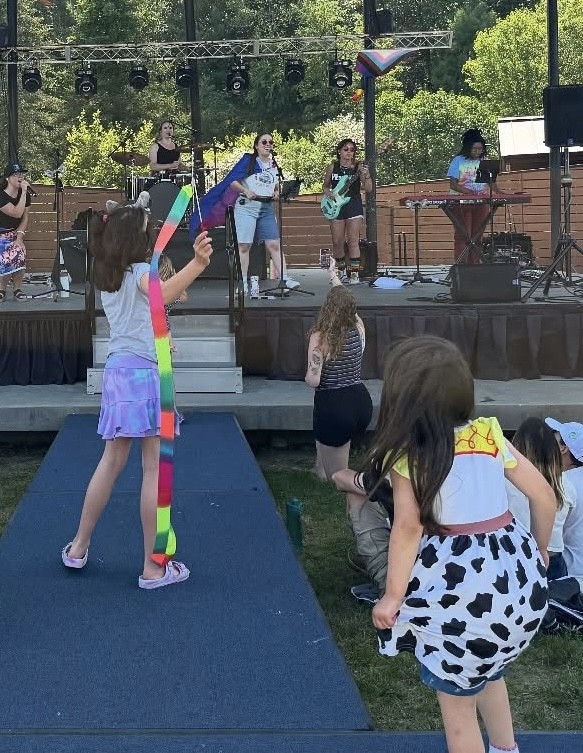 Two young kids, one waving a rainbow streamer and another wearing a cow-printed skirt, dance on the runway in front of the NARA park stage. The band plays in the background.