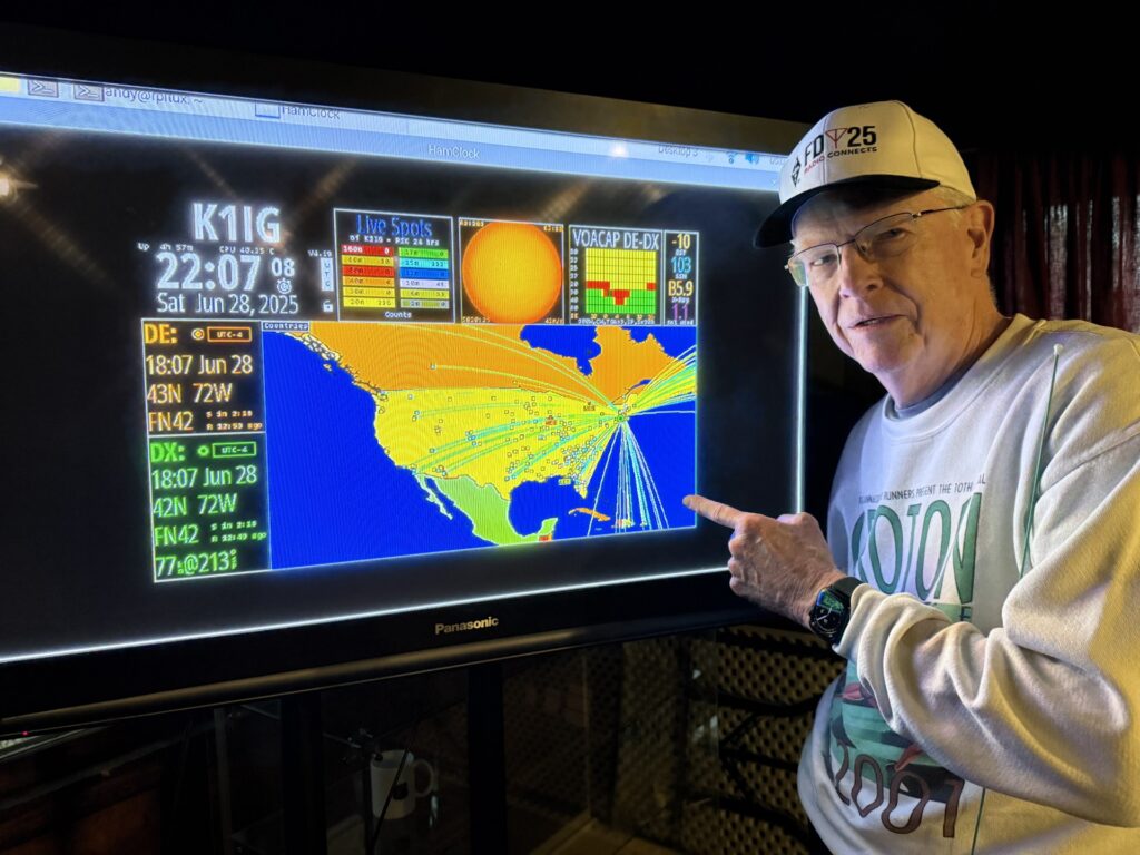 A man in a baseball cap points to a large computer screen. The screen shows a bunch of data and a map with lines radiiating from the northeast (presumably from the Radio Fair location) to locations all over the US, Canada, and points south.