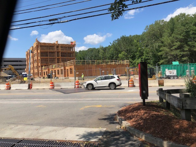 A multi-story wood-framed building in progress. A multi-story parking complex is mostly finished (in the background). Construction equipment, workers, and a wire fence indicate a working construction site.