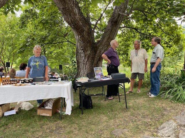 A woman stands behind a table filled with jams and tonics. On the right, a man stands by another table selling tickets for this event.