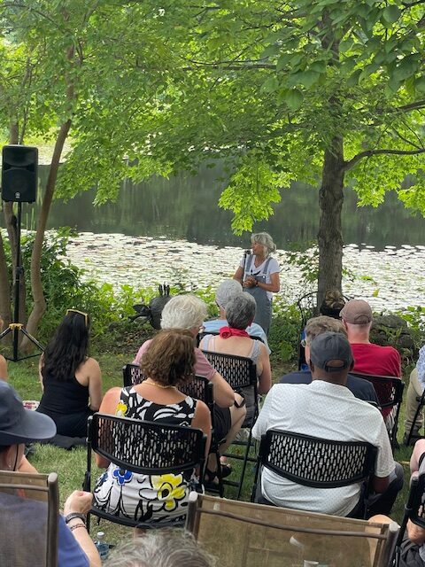 A woman wearing overalls stands under a tree, speaking into a mic. People are seated in front of her, listening.