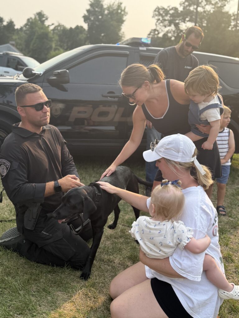 A man sits with a black lab dog. There are several adults petting the dog, in the guise of showing Koda the dog to their young children.