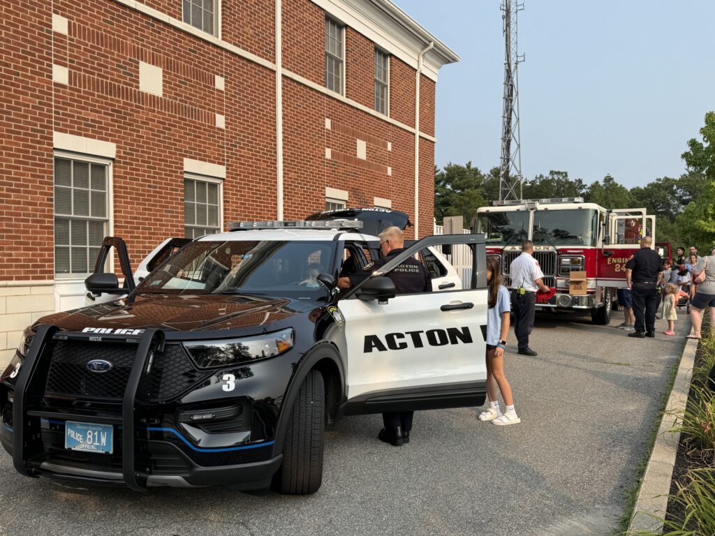 At the side of the building, a police car and fire truck are sitting with their doors open. Kids are checking out the rides.