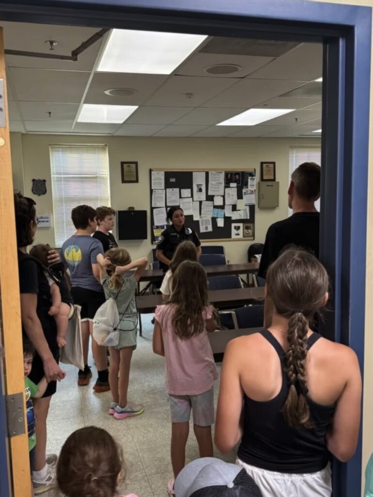 Kids and adults listen to a woman police officer talking inside what looks like a classroom.