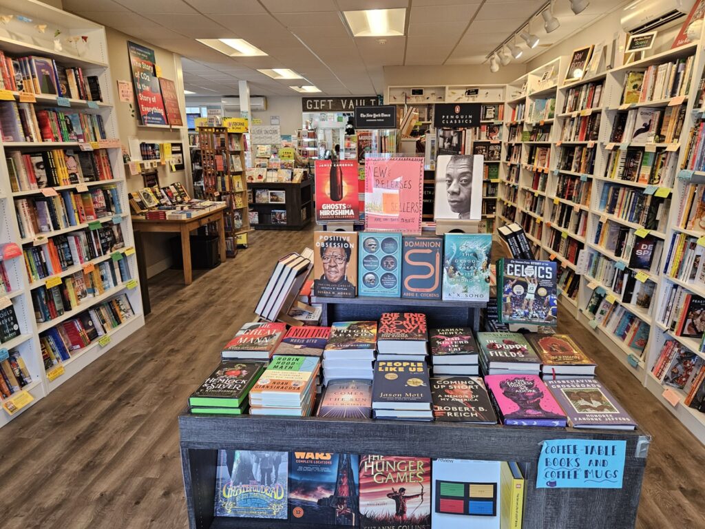 The bookstore from the entrance. On both sides, the walls are lined with books on white bookshelves. In the middle, there are tables full of books.
