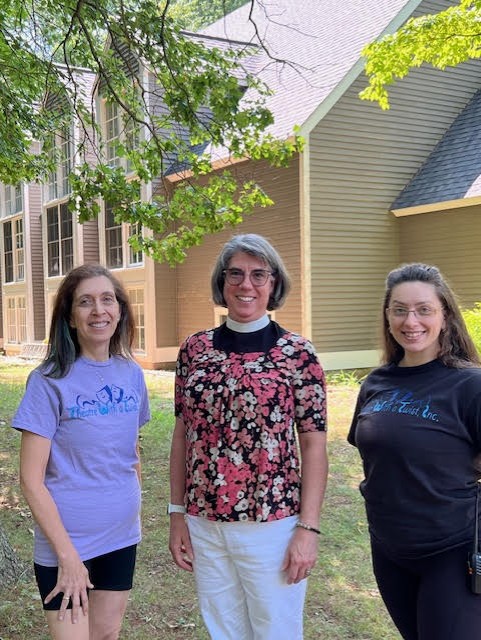 Three women stand in front of a large wooden buildng. The woman in the middle wears a clerical collar and a flowered shirt,