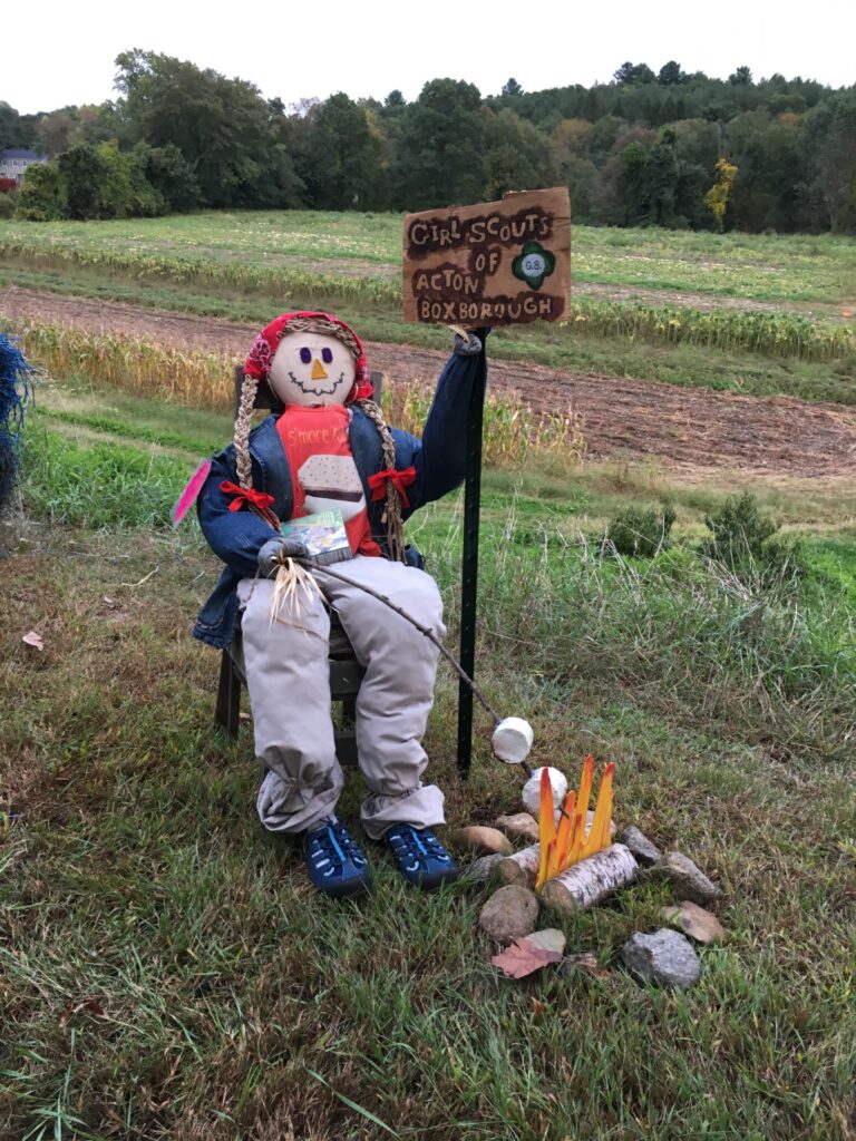 A seated scarecrow dressed as a girl (with braids and a red bandana). She sits in front of a (fake) fire, roasting (fake) marshmallows with one hand. The other hand holds a "Girl Scouts of Acton Boxborough" sign.