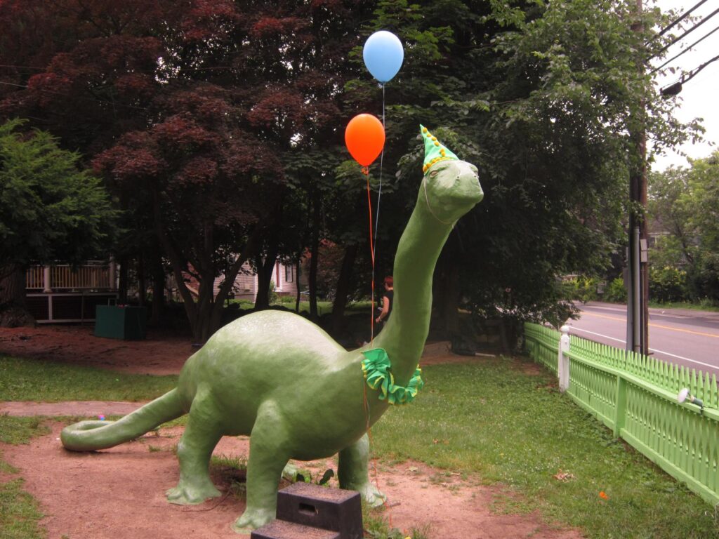 Large, green-painted Triceratops dinosaur sculpture with helium balloons tied to the neck. Green grass, green trees behind it, and a green picket fence bordering the road to the right.