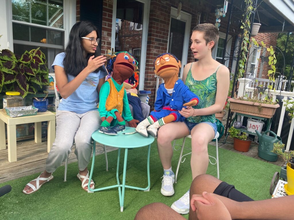 Two young women sit on a sunny patio. One person has a puppet standing on a table next to her, the other has a puppet in her lap.
