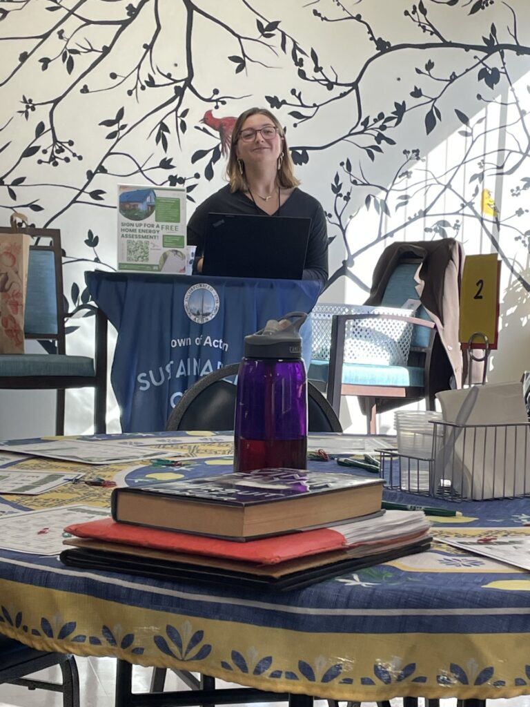 A youngish woman with glasses and shoulder lenght hair stands in front of a wall with tree walpaper. She stands behind a table that has a Town of Acton Sostainibility Office cloth on it.