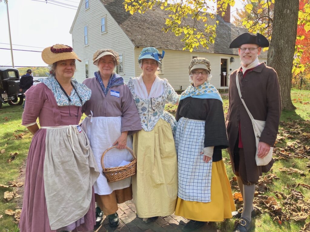 Four women and a man, all in proper Colonial garb, stand in front of an old yellow house on a sunny fall day.